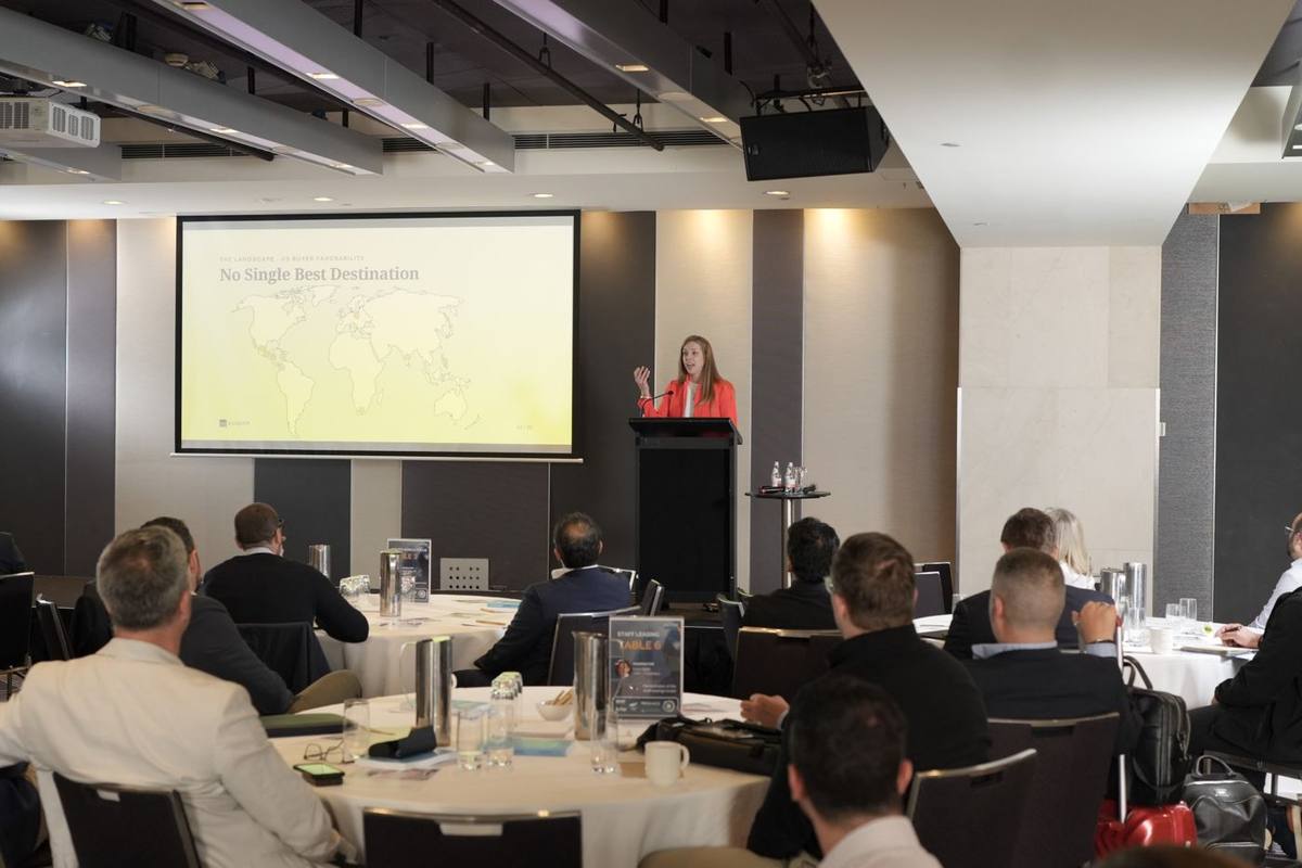 A speaker at a podium presenting to a seated audience at round tables, with a slide on the screen behind her